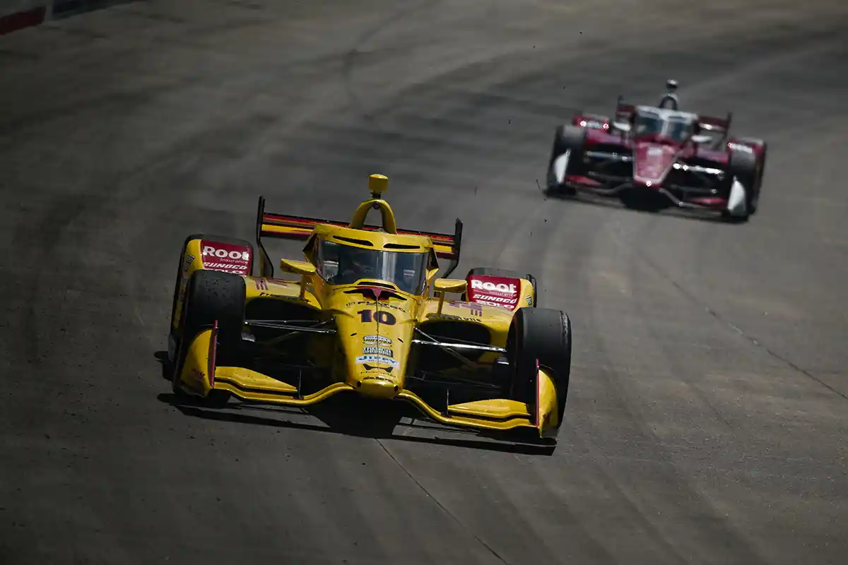 Alex Palou stands beside his IndyCar moments after narrowly missing out on pole position in Portland qualifying, his focused expression contrasting with Felix Rosenqvist’s celebration in the background.