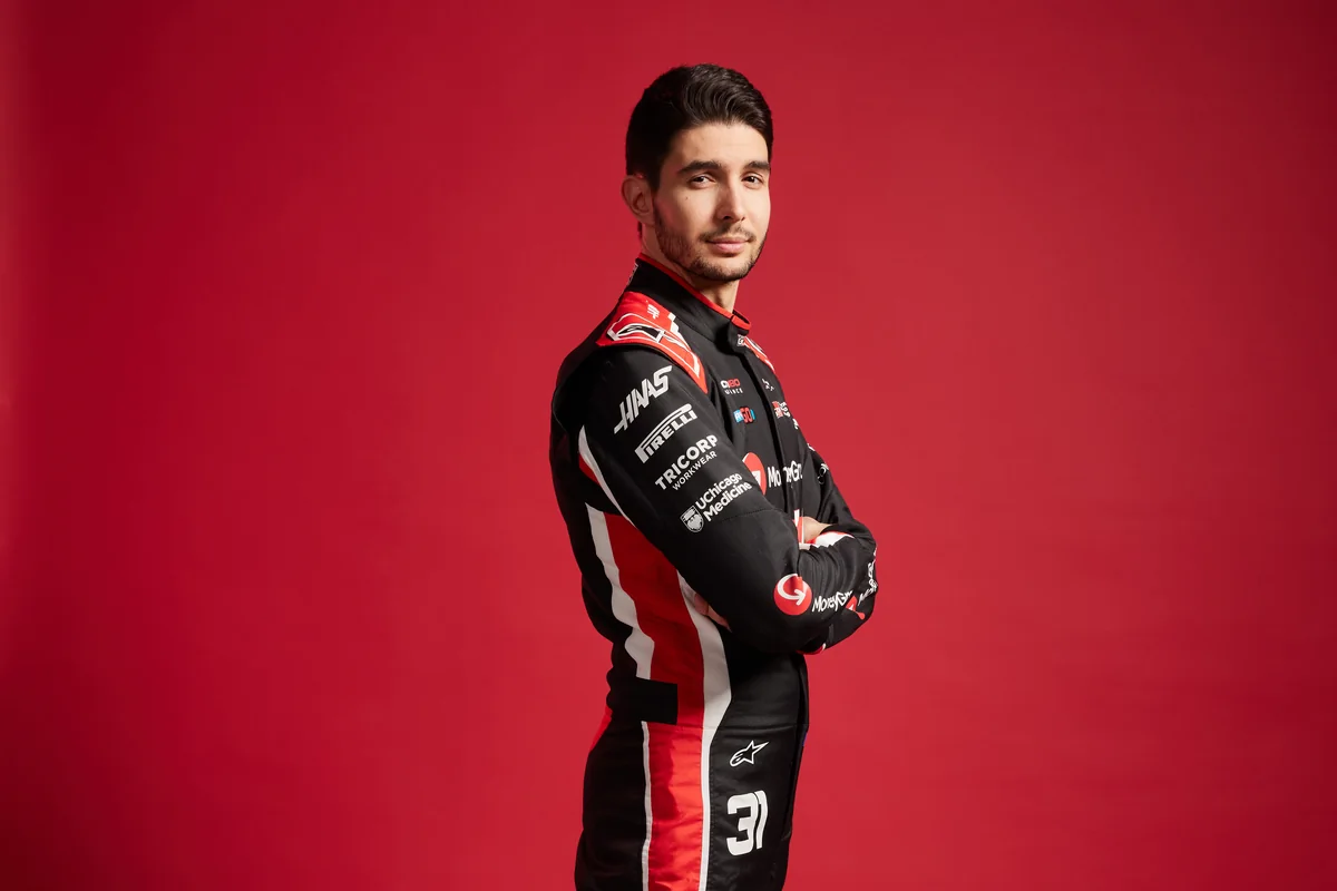 Esteban Ocon in Haas race overalls with his arms folded against a red studio background